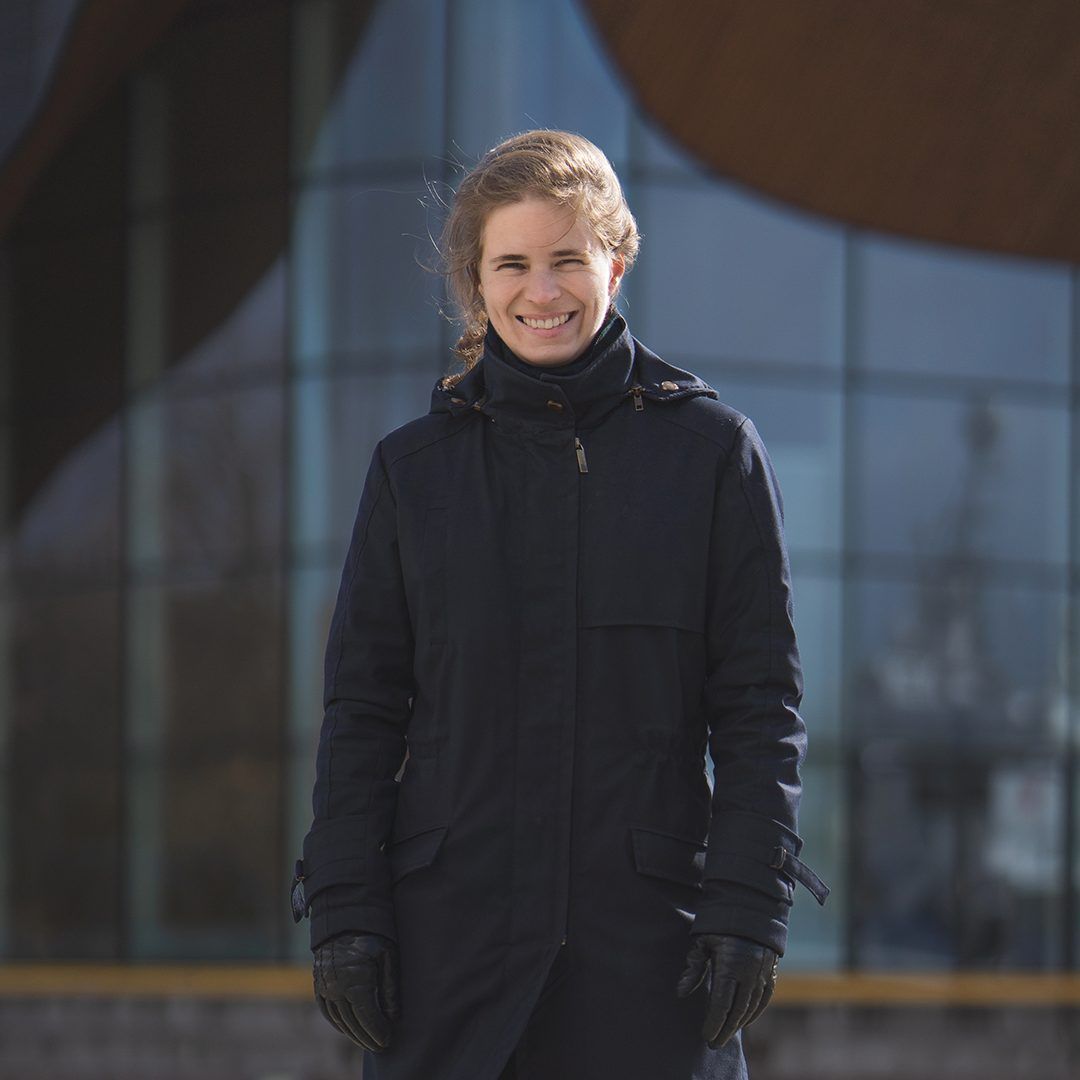 Kvinne med langt mørkt hår som smiler. Hun har på seg en mørk vinterjakke og står foran et bygg med stor glassfasade (Kilden-bygget)