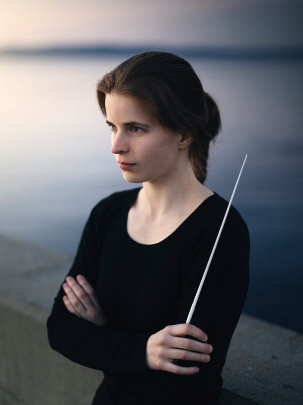 Kvinne med langt mørkt hår som står ved et vann og ser utover. På skuldera har hun en dirigentsokk.