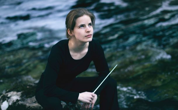 Kvinne som sitter ved et vann med dirigentstokk i hånden og ser opp