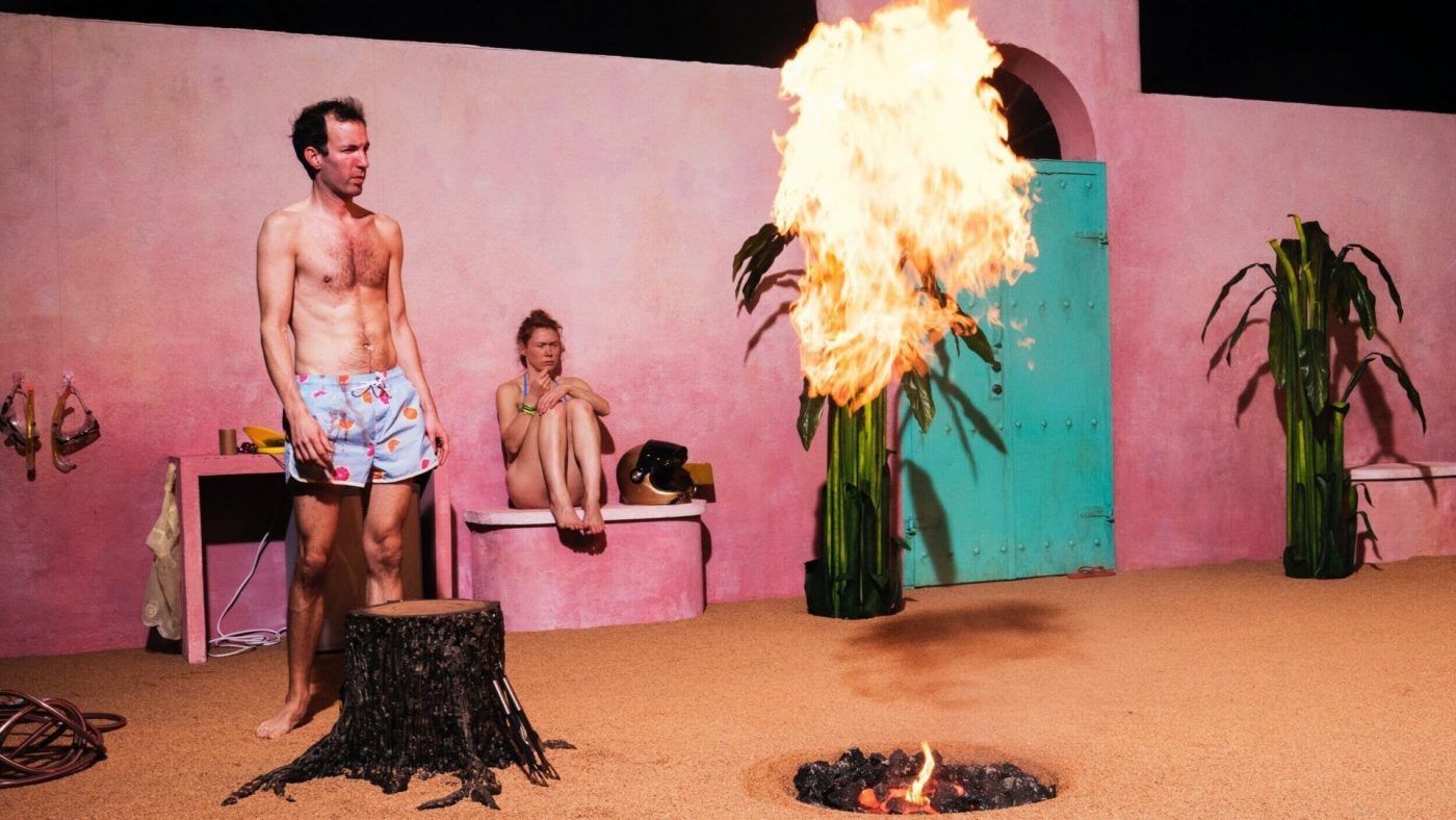 Image from the performance. A man stands in shorts and shirtless looking at a flame rising from the floor. A woman sits on a bench in the background.