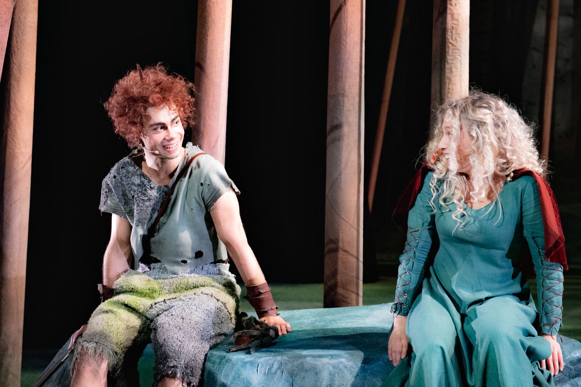 En ung mann og ung kvinne som sitter på en kunstig stein på en scene. De ser på hverandre og smiler.