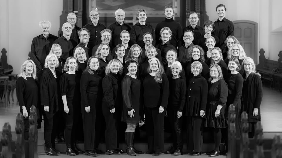 Svart-hvitt gruppebilde av et kor kledd i svart, oppstilt i flere rekker inne i en kirke.